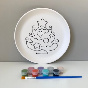 May include: A white ceramic plate with a black outline of a Christmas tree with ornaments. The plate is accompanied by eight small paint pots in various colors and a blue paintbrush.