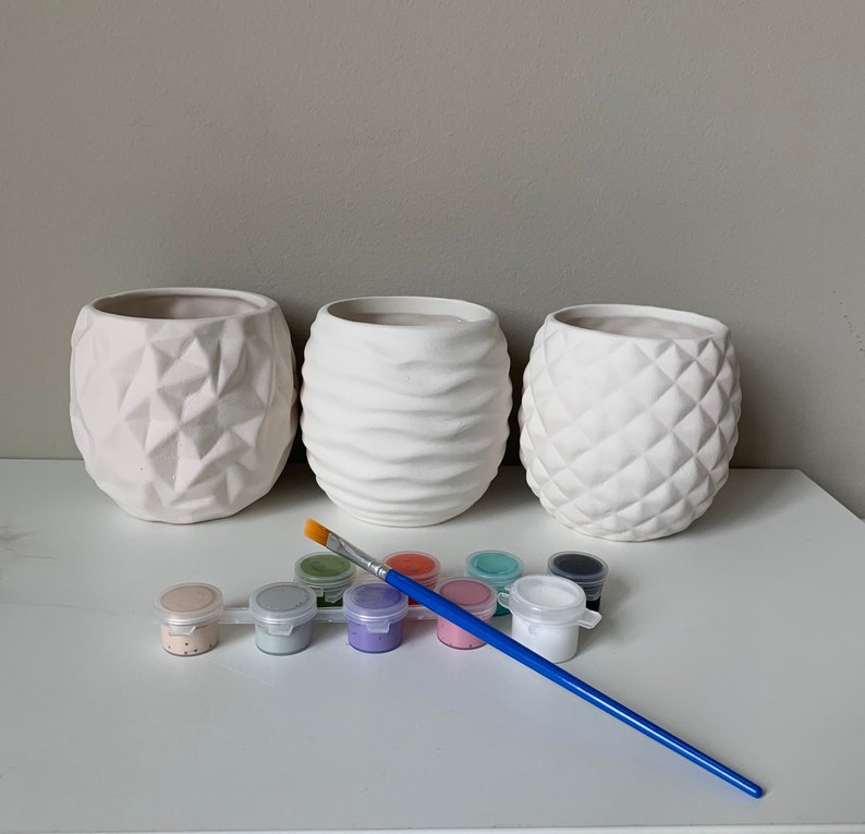 Puede incluir: Tres macetas de cer&aacute;mica blanca con diferentes superficies texturizadas, un pincel y un juego de diez peque&ntilde;os botes de pintura de varios colores.