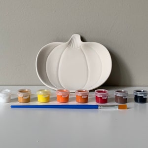 Puede incluir: Un plato de servir de cer&aacute;mica blanco en forma de calabaza con un juego de ocho botes de pintura de varios colores oto&ntilde;ales, incluyendo blanco, marr&oacute;n, amarillo, naranja, rojo y negro, y un pincel azul.
