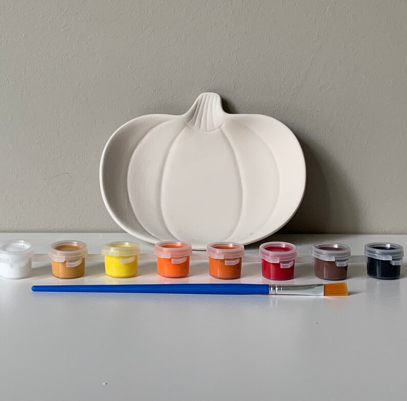 Puede incluir: Un plato de servir de cer&aacute;mica blanco en forma de calabaza con un juego de ocho botes de pintura de varios colores oto&ntilde;ales, incluyendo blanco, marr&oacute;n, amarillo, naranja, rojo y negro, y un pincel azul.