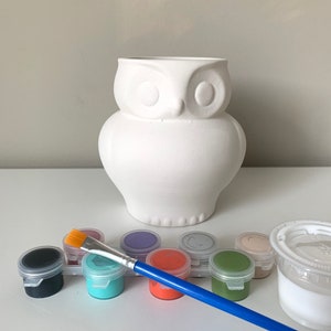 May include: A white ceramic owl-shaped vase with a set of paint pots and a paintbrush. The paint colors include black, teal, orange, and green. The vase is on a white surface.