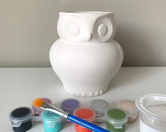 Puede incluir: Un jarrón blanco con forma de búho de cerámica con un juego de botes de pintura y un pincel. Los colores de pintura incluyen negro, turquesa, naranja y verde. El jarrón está sobre una superficie blanca.