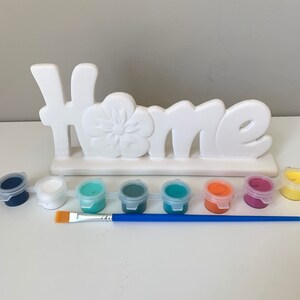 Puede incluir: Un letrero de cer&aacute;mica blanca "Home" con un dise&ntilde;o de flor, listo para ser pintado. El conjunto incluye ocho peque&ntilde;os botes de pintura de varios colores, incluyendo azul, verde, blanco, naranja y rosa, y un pincel azul.