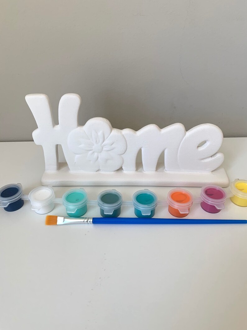 Puede incluir: Un letrero de cer&aacute;mica blanca "Home" con un dise&ntilde;o de flor, listo para ser pintado. El conjunto incluye ocho peque&ntilde;os botes de pintura de varios colores, incluyendo azul, verde, blanco, naranja y rosa, y un pincel azul.