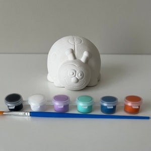 May include: Unpainted ceramic ladybug figurine with six paint pots and a paintbrush. The paint pots are black, white, purple, teal, blue, and orange.