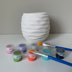 Puede incluir: Un conjunto de botes de pintura de varios colores, incluyendo rosa, morado, verde, naranja, azul y gris. Se incluyen dos pinceles, uno con mango amarillo y otro con mango azul. Los botes de pintura est&aacute;n dispuestos en fila sobre una superficie blanca. Un macetero de cer&aacute;mica blanca con textura ondulada est&aacute; en el fondo.