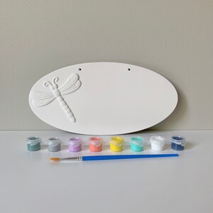 May include: White ceramic oval sign with a dragonfly design, ready to be painted. The set includes eight small paint pots in various colors, including gray, lavender, coral, yellow, turquoise, white, and navy blue, and a paintbrush.