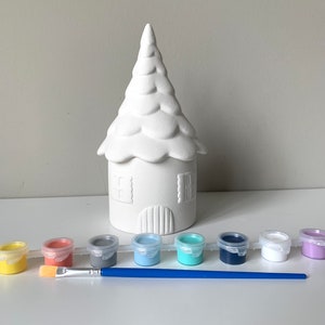 May include: A white ceramic house shaped like a tree with a pointed roof. The house is surrounded by eight small paint pots in various colors, including yellow, orange, gray, light blue, turquoise, navy blue, white, and purple. A blue paintbrush is also included.