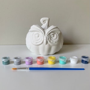 May include: A white ceramic owl pumpkin with a stem, ready to be painted. The set includes eight small paint pots in various colors, including teal, gray, lavender, orange, yellow, turquoise, white, and navy blue, and a blue paintbrush.