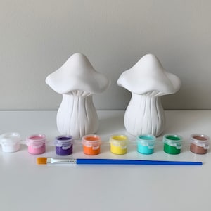 May include: Two white ceramic mushroom figurines with paint pots in various colors, including pink, purple, orange, yellow, turquoise, green, and brown. A blue paintbrush is also included.