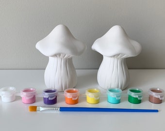 May include: Two white ceramic mushroom figurines with paint pots in various colors, including pink, purple, orange, yellow, turquoise, green, and brown. A blue paintbrush is also included.