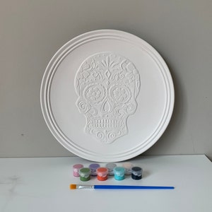 May include: A white ceramic plate with a raised sugar skull design. The plate is surrounded by 10 small paint pots and a blue paintbrush. The paint pots are in various colors, including green, blue, pink, and orange.