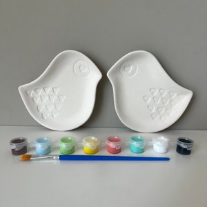 May include: Two white ceramic bird-shaped plates with heart designs, a paintbrush, and eight small paint pots in brown, light blue, green, yellow, coral, turquoise, white, and black.
