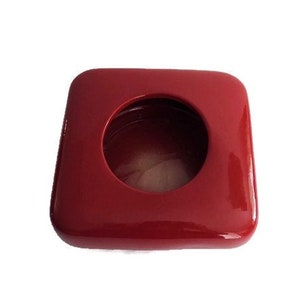 May include: A square, glossy, red container with a circular opening in the center. The container has rounded corners and a smooth, reflective surface. The interior of the opening is a darker shade of red.
