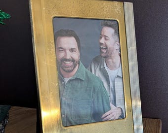 May include: A gold-toned picture frame with a photo of two men smiling. The frame has a textured surface and a silver trim.