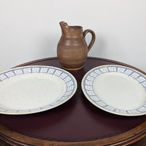 May include: Two white ceramic plates with blue and white patterned borders, one round and one oval, and a brown ceramic jug on a wooden table.