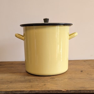 Peut inclure: Une casserole en émail jaune avec un couvercle noir et un bouton noir sur le dessus. La casserole est posée sur une table en bois.