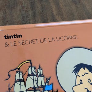 Puede incluir: Una portada de libro con fondo naranja claro que presenta el texto Tintin & LE SECRET DE LA LICORNE en negro. La ilustraci&oacute;n muestra una parte de un velero con velas blancas y una bandera azul, junto a un personaje con un sombrero de ala ancha.