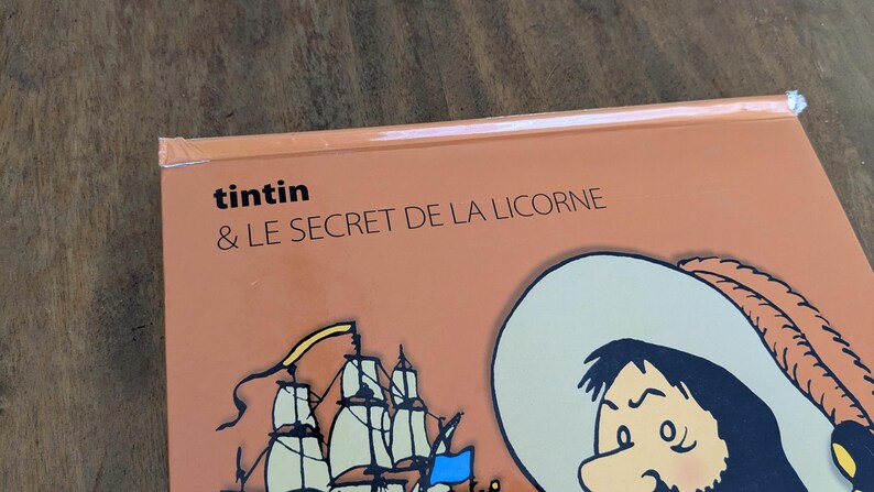 Puede incluir: Una portada de libro con fondo naranja claro que presenta el texto Tintin & LE SECRET DE LA LICORNE en negro. La ilustraci&oacute;n muestra una parte de un velero con velas blancas y una bandera azul, junto a un personaje con un sombrero de ala ancha.