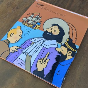 Puede incluir: Una portada de libro titulada Tintin & Le Secret de la Licorne con una colorida ilustraci&oacute;n de Tint&iacute;n, el Capit&aacute;n Haddock y una figura hist&oacute;rica con sombrero de ala ancha, con un velero de fondo sobre un cielo azul claro.