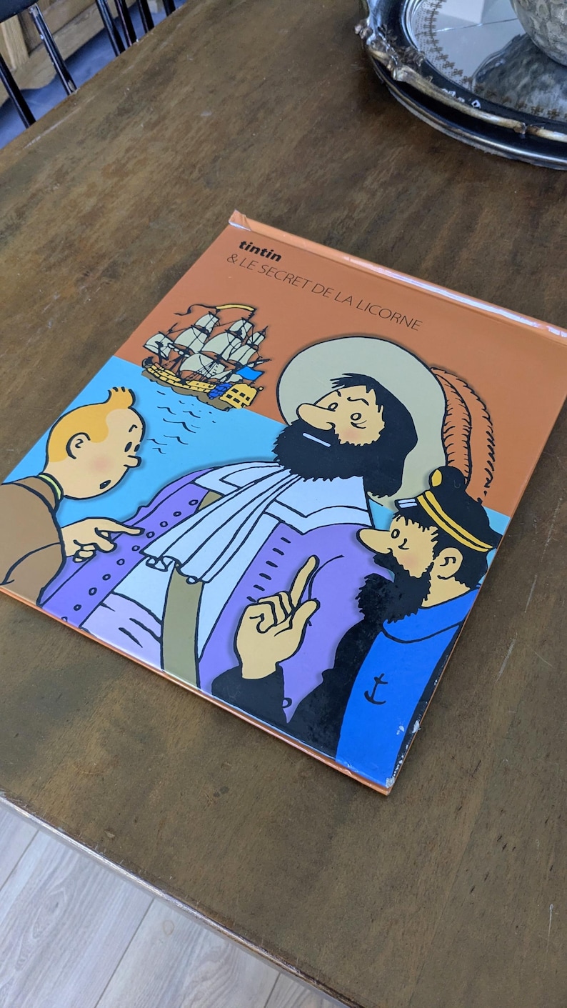 Puede incluir: Una portada de libro titulada Tintin & Le Secret de la Licorne con una colorida ilustraci&oacute;n de Tint&iacute;n, el Capit&aacute;n Haddock y una figura hist&oacute;rica con sombrero de ala ancha, con un velero de fondo sobre un cielo azul claro.