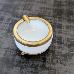 May include: A small, round ashtray with a white interior and a gold-colored rim and accents. The ashtray has a small rest for cigarettes and sits on three small feet. A classic smoking accessory.