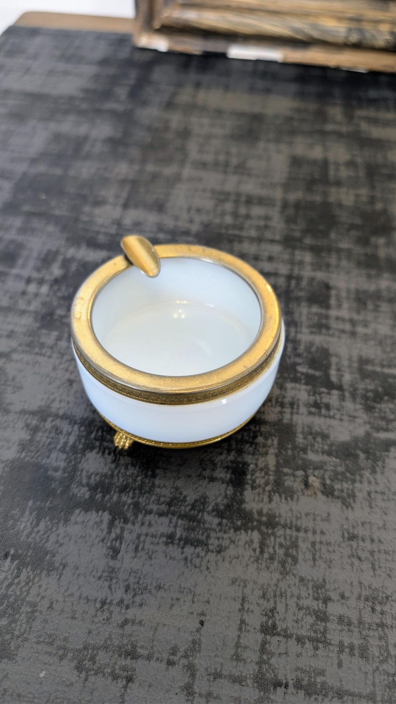 May include: A small, round ashtray with a white interior and a gold-colored rim and accents. The ashtray has a small rest for cigarettes and sits on three small feet. A classic smoking accessory.