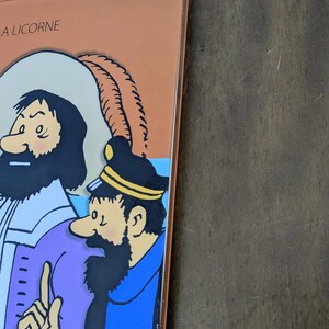 Puede incluir: Una portada de libro de tapa dura con una ilustraci&oacute;n de dibujos animados de dos personajes, uno con abrigo morado y otro con uniforme azul con un emblema de ancla. El texto "A LICORNE" est&aacute; impreso en negro en la esquina superior izquierda.