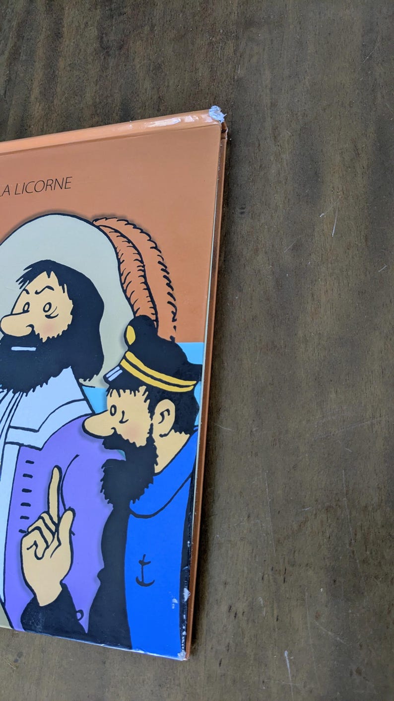 Puede incluir: Una portada de libro de tapa dura con una ilustraci&oacute;n de dibujos animados de dos personajes, uno con abrigo morado y otro con uniforme azul con un emblema de ancla. El texto "A LICORNE" est&aacute; impreso en negro en la esquina superior izquierda.
