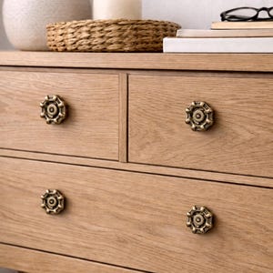 Könnte beinhalten: Eine helle Eichenkommode mit vier Schubladen, jede mit einem dekorativen Messingknauf im antiken Stil. Auf der Kommode befinden sich eine Vase, ein geflochtener Korb, eine Kerze und Bücher. Die Holzmaserung ist sichtbar.