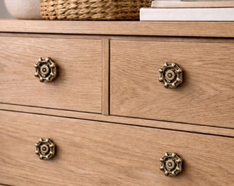 May include: A light oak dresser with four drawers, each featuring a decorative, antique-style brass knob. The dresser top holds a vase, a woven basket, a candle, and books. The wood grain is visible.