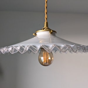May include: A white glass pendant light fixture with a scalloped edge and a gold metal base. The light fixture is hanging from a braided cord and has a clear light bulb.