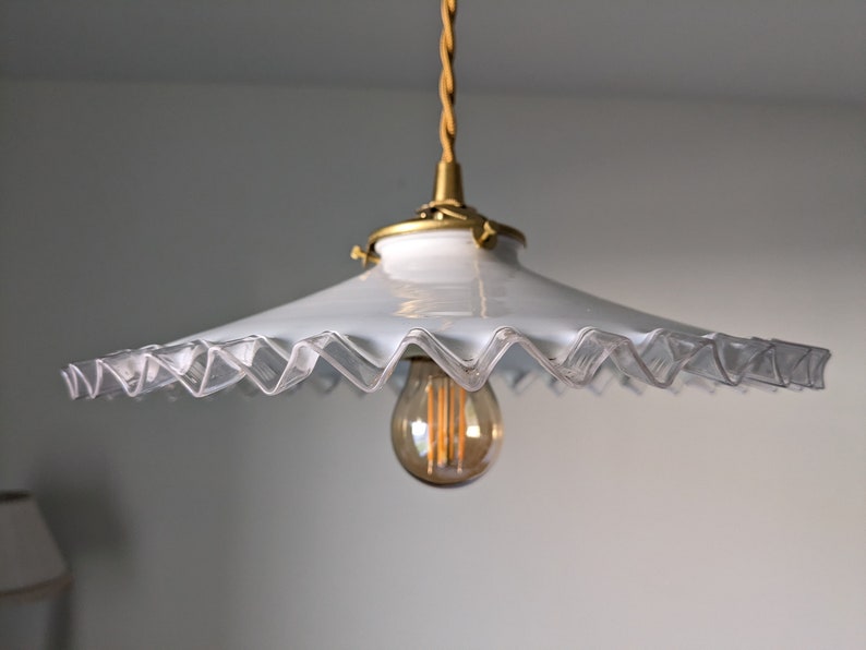 May include: A white glass pendant light fixture with a scalloped edge and a gold metal base. The light fixture is hanging from a braided cord and has a clear light bulb.