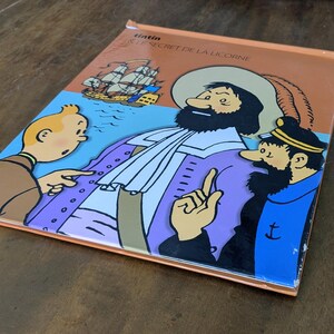 Puede incluir: Un c&oacute;mic de Tint&iacute;n titulado Le Secret de la Licorne con una colorida ilustraci&oacute;n de Tint&iacute;n, el Capit&aacute;n Haddock y una figura hist&oacute;rica con abrigo morado. La portada incluye un barco de vela sobre un fondo azul y bordes de color naranja.