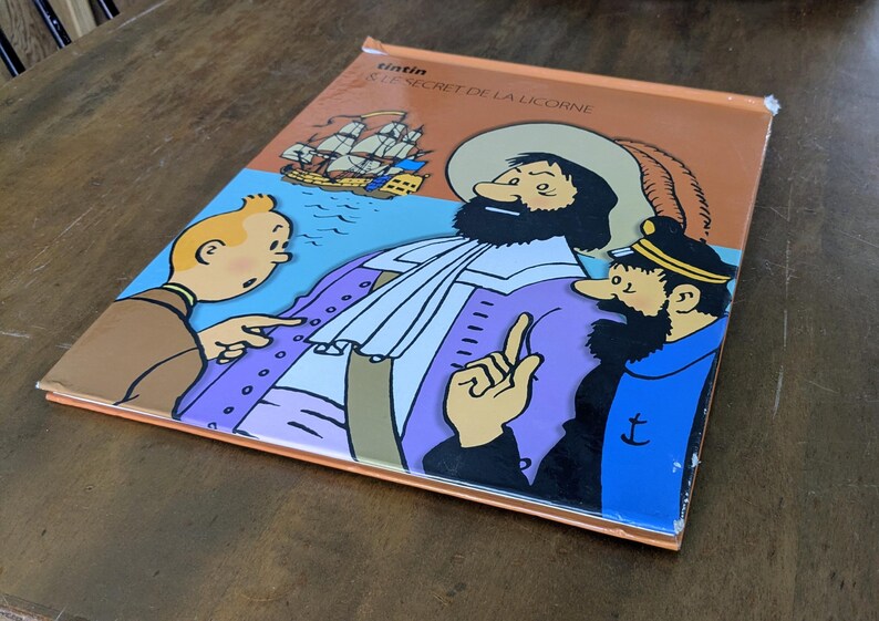 Puede incluir: Un c&oacute;mic de Tint&iacute;n titulado Le Secret de la Licorne con una colorida ilustraci&oacute;n de Tint&iacute;n, el Capit&aacute;n Haddock y una figura hist&oacute;rica con abrigo morado. La portada incluye un barco de vela sobre un fondo azul y bordes de color naranja.