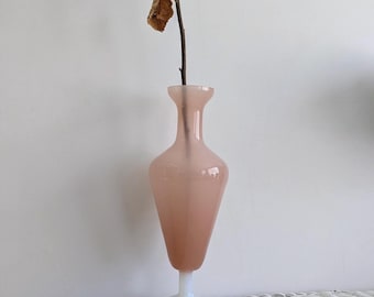 May include: A pale pink glass vase with a slender neck and a tapered body, supported by a solid white circular base. A single thin brown stem with a few dried, withered brown leaves extends vertically from the opening of the glass vessel.
