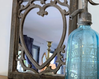 Op de afbeelding: Decoratieve spiegel met een hartvormige opening en een sierlijk houten frame. Een vintage blauwe glazen sodasifonfles staat rechts. Een messing kandelaar wordt weerspiegeld in de spiegel.