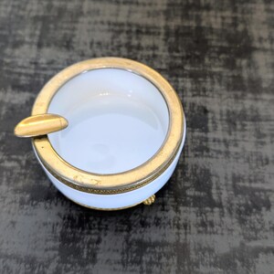 May include: A small, round ashtray with a white interior and a gold-colored rim and cigarette rest. The ashtray has three small gold-colored feet. The ashtray is on a dark surface.
