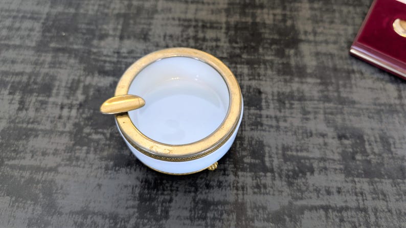 May include: A small, round ashtray with a white interior and a gold-colored rim and cigarette rest. The ashtray has three small gold-colored feet. The ashtray is on a dark surface.