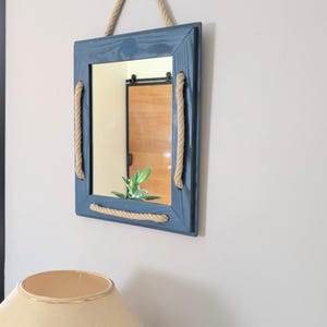 Op de afbeelding: Een rechthoekige blauwe spiegel met touwdetails. De spiegel is omlijst in blauw hout en heeft touwaccenten aan de zijkanten en bovenkant om op te hangen. De spiegel weerspiegelt een kamer met een houten deur en een plant.