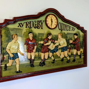 Peut inclure: Plaque murale décorative en bois sur le thème du rugby, avec une horloge. Le cadre est rouge foncé et porte l'inscription "XV Rugby Quinze XV". L'horloge indique environ 3h00.