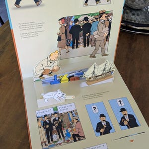 Puede incluir: Un libro desplegable con ilustraciones de Tint&iacute;n y texto en franc&eacute;s. La p&aacute;gina muestra una escena en 3D con un modelo de barco en miniatura, una figura de Tint&iacute;n agachado y un perro blanco. Paneles incluyen c&oacute;mics con personajes.