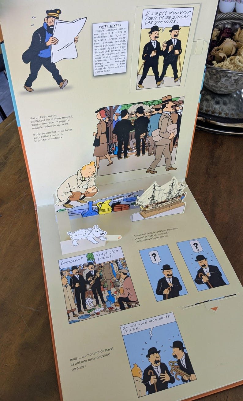 Puede incluir: Un libro desplegable con ilustraciones de Tint&iacute;n y texto en franc&eacute;s. La p&aacute;gina muestra una escena en 3D con un modelo de barco en miniatura, una figura de Tint&iacute;n agachado y un perro blanco. Paneles incluyen c&oacute;mics con personajes.