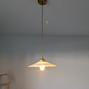 May include: A white, scalloped-edge lampshade hangs from a gold cord and ceiling fixture. The lampshade is illuminated by a single light bulb.