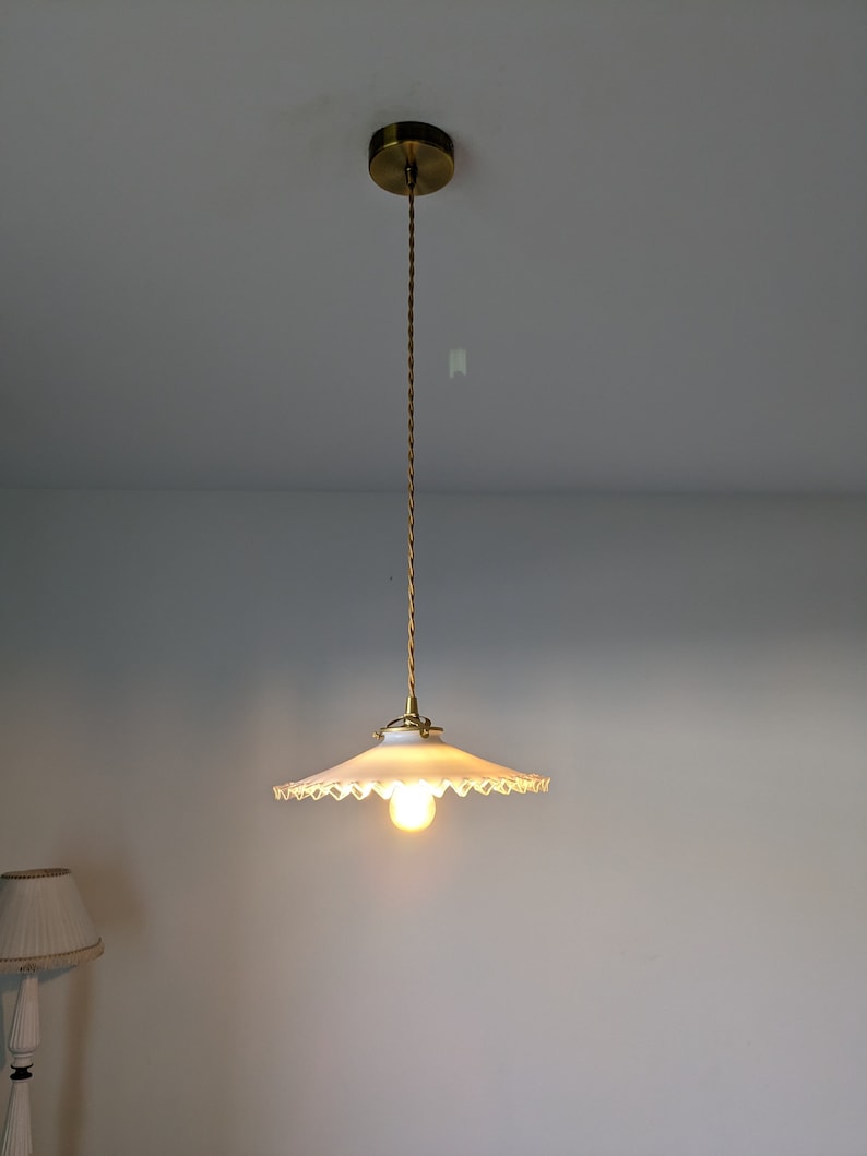 May include: A white, scalloped-edge lampshade hangs from a gold cord and ceiling fixture. The lampshade is illuminated by a single light bulb.