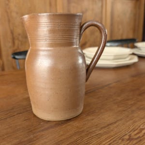 Op de afbeelding: Een bruine steengoed kruik met een gebogen handvat. De kruik heeft een brede basis en loopt taps toe naar de bovenkant, met horizontale ribbels bij de tuit. Hij staat op een houten tafel.