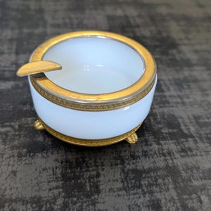 May include: A white and gold ashtray with a circular design. The ashtray has a gold rim and a small rest for a cigarette. The ashtray sits on three small gold feet.