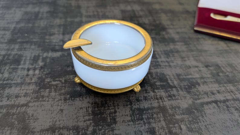 May include: A white and gold ashtray with a circular design. The ashtray has a gold rim and a small rest for a cigarette. The ashtray sits on three small gold feet.