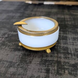 May include: A white and gold ashtray with a round shape and three small feet. The ashtray has a gold rim and a small rest for cigarettes. The ashtray is made of ceramic and metal.