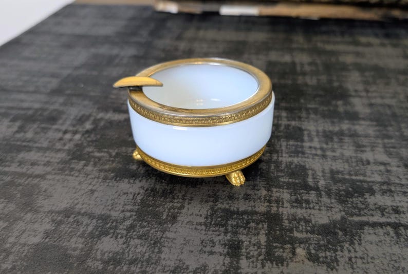 May include: A white and gold ashtray with a round shape and three small feet. The ashtray has a gold rim and a small rest for cigarettes. The ashtray is made of ceramic and metal.